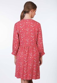 Rotes Blumenkleid mit Dreiviertelärmeln, geraffter Taille und einem Muster, das kleine weiße und gelbe Blumen zeigt. Glatte Stoffoberfläche.