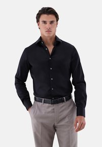 Schwarzes langärmeliges Hemd mit Knöpfen und spitzem Kragen, kombiniert mit hellgrauen Hosen. Aus glattem Stoff mit dezenten Knöpfen.