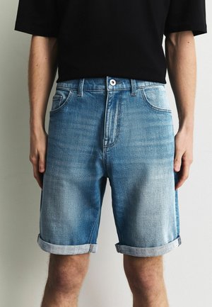 Férfi világoskék, felhajtott farmer rövidnadrágban és fekete rövid ujjú ingben, egyszínű háttér előtt állva.