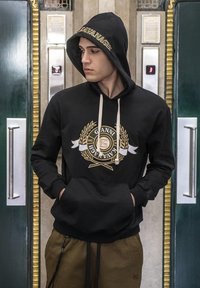Sudadera negra con un logo dorado que presenta hojas de laurel y texto. Incluye cordones beige y un bolsillo frontal. Estilizada contra un fondo interior.