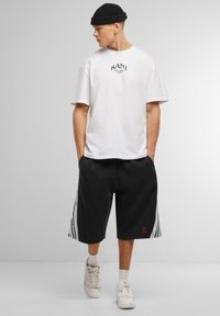 White cotton t-shirt with printed logo, paired with black shorts featuring white stripe patterns and red brand detail. White sneakers.