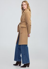 Manteau en mélange de laine beige avec une taille à liens, col à revers, poches latérales, associé à un jean bleu à jambes larges et des bottines à talons noires. 