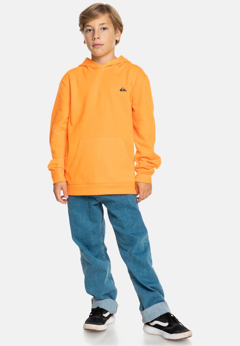 Sudadera naranja brillante con un bolsillo frontal, combinada con pantalones vaqueros azul claro y zapatos negros con suelas blancas. Diseño simple, sin patrones.
