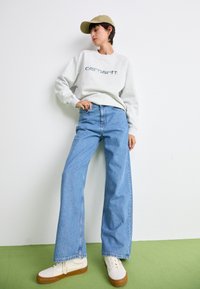 Gray sweatshirt with "Carhartt" logo, blue wide-leg jeans, and white sneakers with brown soles, complemented by a light green background.
