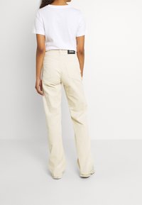 Person, der beige Cordhosen und ein weißes T-Shirt trägt, steht mit dem Rücken zu einer schlichten weißen Hintergrund.