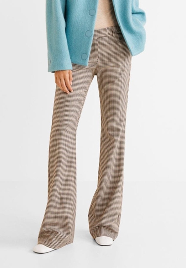 Femme portant un pantalon large marron à carreaux, une veste bleu clair boutonnée et des chaussures blanches, debout sur un fond blanc.