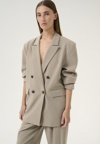 Beige, doppelt geknöpfte Blazer mit tiefem V-Ausschnitt, drei Viertelärmeln und schwarzen Knöpfen; glattes Material, strukturierte Passform und seitliche Taschen.