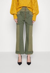 Knopvoorkant groene broek met wijd uitlopende pijpen, contrasterende panelen en grote zakken aan de voorkant. Het model combineert ze met een gestructureerde gele trui en zwarte hakken.