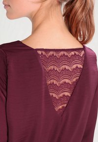 Burgundy topp med djup V-ringning, prydd med ett fint spetsinlägg med en vågig mönstring. Slät textur, minimalistisk design.