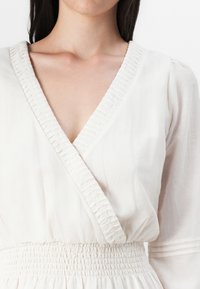 Primo piano della parte superiore del corpo di una persona in un vestito bianco morbido con scollatura a volant e vita smock, posto su uno sfondo semplice.