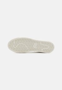 Fehér sneaker talp hullámos mintázattal, texturált felülettel és logólenyomattal a sarkán. A talp kialakítása javítja a tapadást.