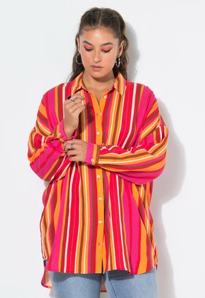 Junge Frau mit langen dunklen Haaren trägt ein übergroßes Hemd mit leuchtenden vertikalen Streifen in Orange, Pink, Rot und Gelb und richtet ihren Ärmel.