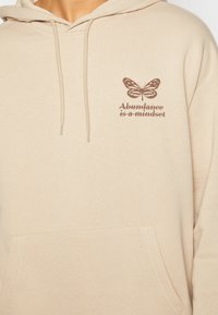 Felpa in misto cotone beige con tasca frontale, cappuccio con cordino e stampa di una farfalla marrone con testo sul petto.