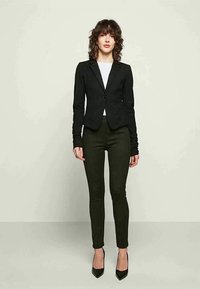 Schwarzes tailliertes Sakko mit einem einzigen Knopf, kombiniert mit olivgrünen Slim-Fit-Hosen und schwarzen High Heels. Glattes Material, strukturierte Form.