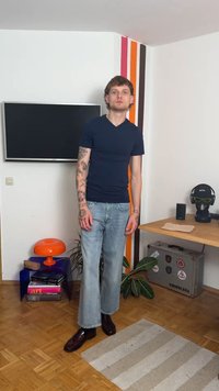 Jūras spēku pieguļošs T-krekls, gaiši zili plata kāju džinsi, brūni eleganti apavi. Redzami tetovējumi uz rokām. Vienkārša istaba ar mēbelēm un dekoriem.