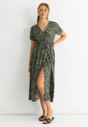 Woman wearing a green leopard print wrap midi dress with a side slit and black slide sandals, standing against a white background.
