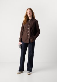 Brown button-up shirt made of cotton, paired with dark denim wide-leg pants. Model holds a silver clutch and wears grey shoes.