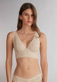 Lace bralette in light beige with a V-neck design, featuring intricate floral patterns and a small bow detail at the center front.