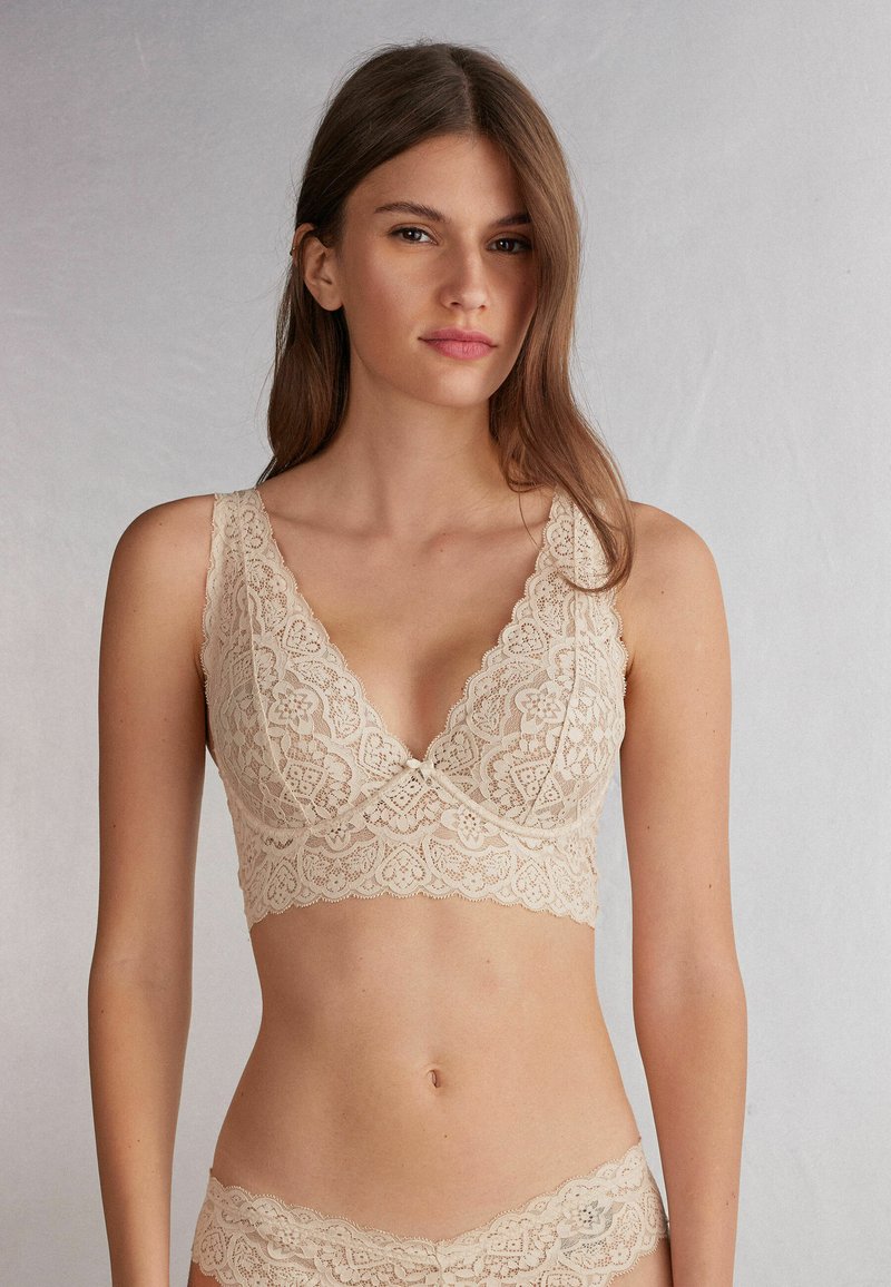 Lace bralette in light beige with a V-neck design, featuring intricate floral patterns and a small bow detail at the center front.