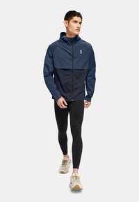 Lightweight navy jacket with a contrasting dark navy section, zip front, and a hood, paired with black leggings and white running shoes.