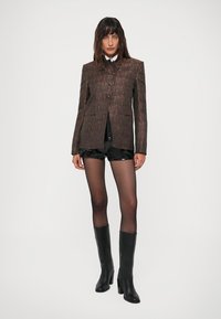Femeie îmbrăcată cu un sacou întunecat cu model, pantaloni scurți negri, ciorapi negri translucizi și cizme negre până la genunchi, stand în fața unui fundal simplu.