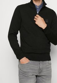 Svart tröja med dragkedja i halsen, stickad textur, ribbade ärmslut och nederkant, bärs över en blåmönstrad skjorta, händerna placerade avslappnat.