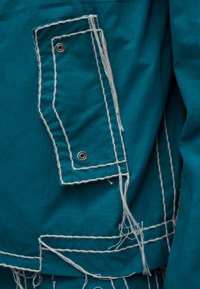 Πάνελ από πετροπράσινο ύφασμα με μεγάλες ραμμένες λευκές γραμμές, μεταλλικά κουμπιά με πάτημα και χαλαρές άκρες νημάτων που δημιουργούν μια υφασμάτινη, πολυστρωματική λεπτομέρεια ρούχου.