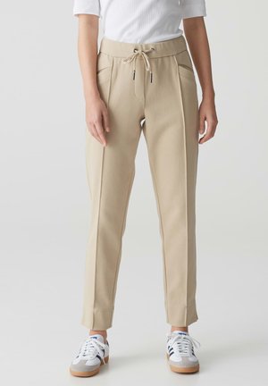 Beiges Jogger-Hosen mit elastischem Bund, seitlichen Taschen, schmal zulaufendem Bein und glatter Textur, kombiniert mit weißen und grauen Sneakers.