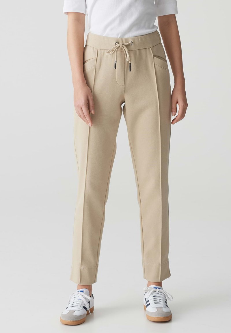 Beige joggerbroeken met een trekkoord in de taille, zijzakken, taps toelopende pijpen en een soepele textuur, gecombineerd met wit-grijze sneakers.