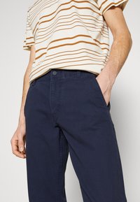 Kortärmad t-shirt med rund halsringning i tunna bruna och krämfärgade ränder, kombinerad med marinblå byxor med framfickor och knäppning.