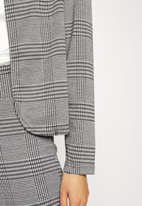 Blazer à motif pied-de-poule noir et blanc, doté d'un col cranté, de manches longues et de bords nets, fabriqué en tissu tissé.