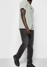 Camiseta de algodón verde claro con un logo en el pecho, combinada con unos vaqueros oscuros gris y zapatillas negras. El conjunto presenta un corte relajado y un estilo casual.