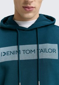 Teal hoodie met een grafisch logo in wit en donkerblauw. Gemaakt van een zachte stof met trekkoorden en een kangoeroezak.