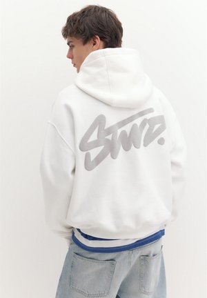 Junger Mann trägt einen weißen Hoodie mit großem stilisiertem Text auf dem Rücken, hellblaue Jeans und ein blau-weiß gestreiftes Hemd darunter.