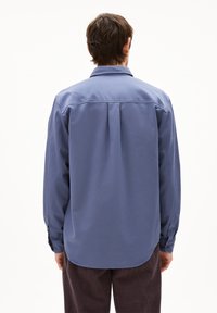 Camicia a maniche lunghe blu chiaro con colletto reversibile, piega sul retro e polsini. Presenta una texture liscia e un orlo arrotondato.