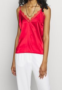 Rød satin camisole med V-udskæring og blonder, tynde justerbare stropper og en glat tekstur. Sættes sammen med hvide bukser.