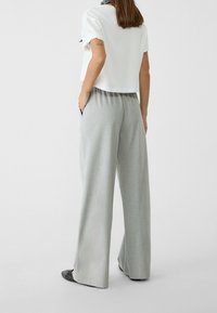 Kobieta w jasnoszarych szerokich spodniach, białej krótkiej bluzce z krótkim rękawem, czarnych butach i czarno-białym szaliku na szyi, stoi z rękami w kieszeniach.