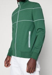 Giacca verde con zip e strisce bianche, realizzata in tessuto morbido. Presenta tasche laterali con zip e un colletto alto. Polsini e orlo elastici.