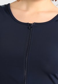 Costume da bagno blu navy con scollo rotondo e chiusura con cerniera frontale. Realizzato in un tessuto liscio con un design aderente. Dettagli testurizzati lungo la cerniera.
