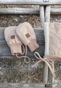 Vaaleanpunaiset lapaset, jotka on valmistettu pehmeästä kankaasta ja joissa on ribbireuna ja logopatchi, ovat vieressä vaaleanpunainen turkikseen vuorattu asuste, jossa on sidottu nauha.