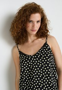 Camisola in satin nero con pois bianchi e verdi, bordo in pizzo lungo il colletto, spalline sottili regolabili, vestibilità rilassata, texture liscia.