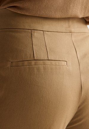 Hnědé kalhoty na míru s jemnou diagonální texturou, vybavené zadní kapsou a čistými švy, zdůrazňujícími hladký povrch a tapered střih.