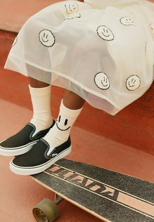 Persona che indossa calzini bianchi con faccine sorridenti, scarpe slip-on nere e una gonna trasparente con motivi di faccine sorridenti, seduta sopra uno skateboard.