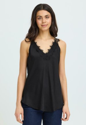 Mladá žena s dlhou tmavou vlasmi, ktorá má na sebe čierny bezrukávový top s čipkovým lemom a modré džínsy, stojaca pred jednoduchým pozadím.