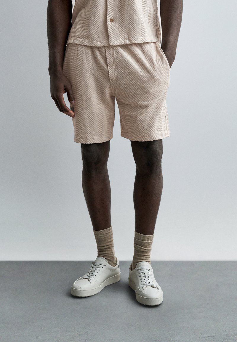 Homme portant un short à motif chevrons beige, un haut assorti, des chaussettes beige et des baskets blanches basses, debout sur un sol gris.