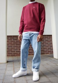 Felpa bordeaux con collo a giro, logo bianco, jeans in denim blu con risvolti, e sneaker bianche con dettagli texturizzati sulle suole.