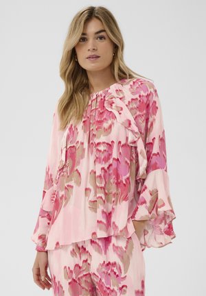 Donna con lunghi capelli biondi che indossa una camicetta astratta floreale rosa e beige con maniche arricciate, in piedi davanti a uno sfondo grigio semplice.