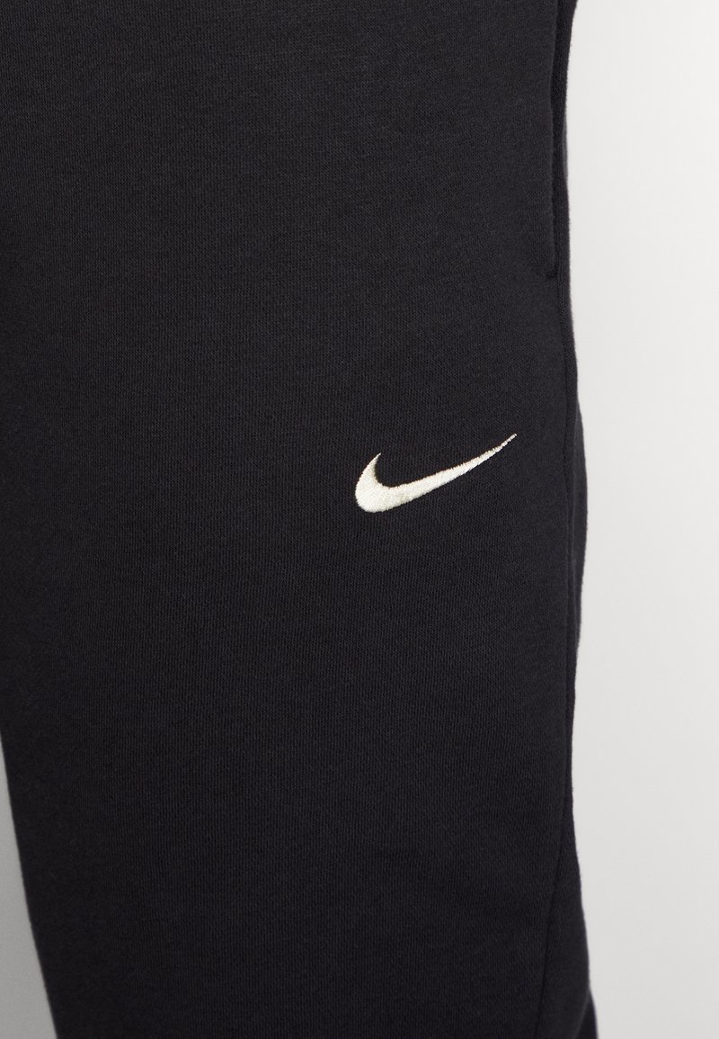 Pantalones jogger negros hechos de una tela suave y texturizada. Presenta un logo blanco de Nike en el muslo izquierdo y bolsillos laterales. Diseño simple y atlético.