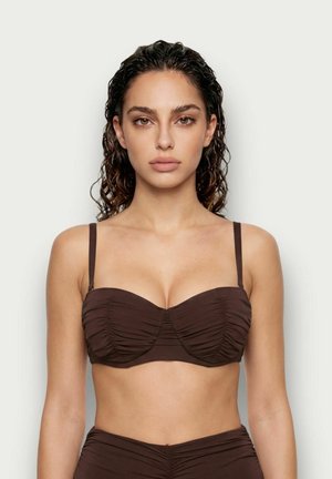 Femme aux cheveux bouclés mouillés portant un haut de bikini froncé marron foncé, face à un fond clair uni.