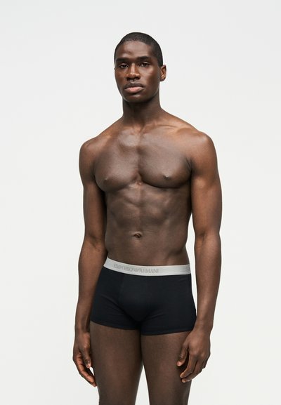 Jämes lihaseline mees seisab, kandes musti Emporio Armani bokserpükse halli vöökohaga, lihtsa heleda tausta ees.